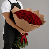 Person holding a large bouquet of red roses wrapped in brown paper with a red ribbon against a gray background