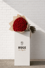 Bouquet of red roses wrapped in brown paper with 'HUGE Los Angeles' branding against a white brick wall.