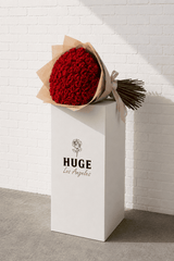 Bouquet of red roses in brown paper packaging on a white pedestal with 'HUGE Los Angeles' branding against a white brick wall.
