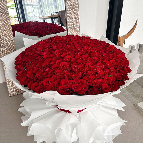 Large bouquet of red roses wrapped in white paper with a silk ribbon, displayed in an interior setting.